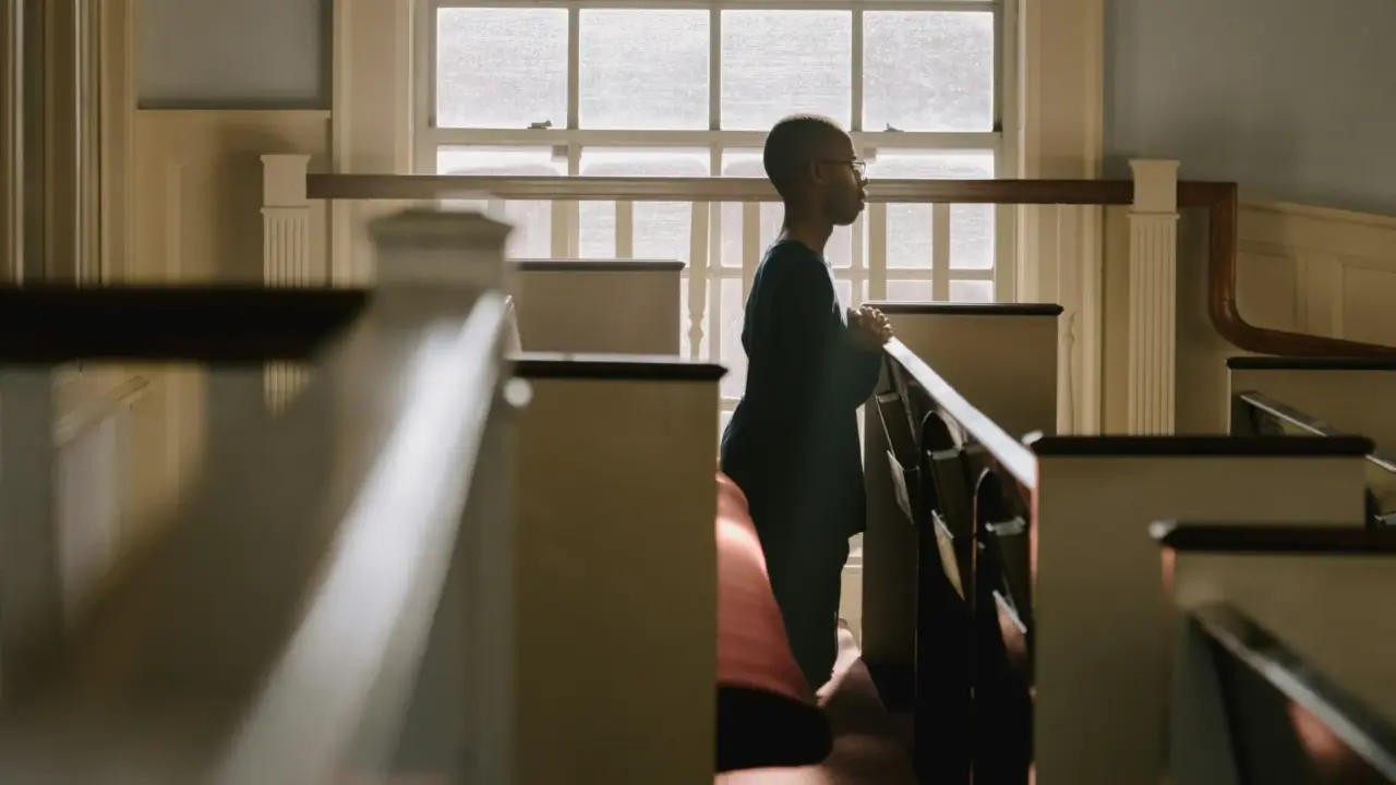 Man kneeling and praying over church pew beside window | Religious Trauma | Elnita Ottey & Associates